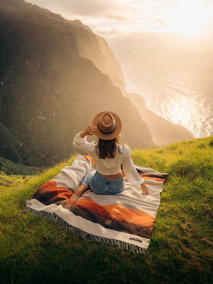 A woman enjoying the scenic view of the ocean while sitting on a blanket on a hill.