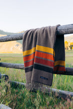 Load image into Gallery viewer, A cozy blanket hanging on a fence in a beautiful field.
